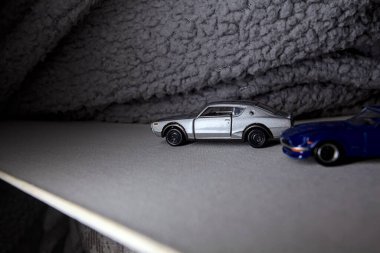 Mantova, İtalya - 3RD JANUARY 2025 Takara Tomy kopyaları gri Nissan Skyline 2000 GT-R ve mavi Nissan Fairlady Z beyaz bir panoda, gri bir kumaş parçasının yanında, uçurumun kenarındaki bir dağ yolunda yarış yapıyormuş gibi yapıyor.