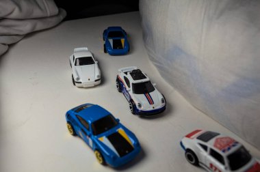 Mantova, İtalya - 4. JANUARY 2025 - çeşitli Porsche arabalarının Hot Wheels kopyaları, bir 911 rallisi, iki 1996 Carrera, bir 1971 911 ve bir 911 Carrera RS 2.7, beyaz tahta üzerinde, beyaz kumaşlarla çevrili, yolda bir eğri yapıyormuş gibi yaparak