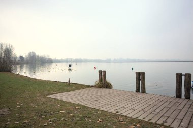 Göl kıyısında ahşap demirleme direkleri olan iskele ve İtalya kırsalında bulutlu ve sisli bir günde suya dökülen arka plandaki ağaçlar.
