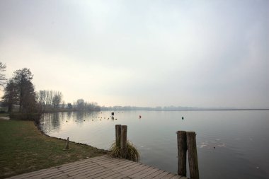 Göl kıyısında ahşap demirleme direkleri olan iskele ve İtalya kırsalında bulutlu ve sisli bir günde suya dökülen arka plandaki ağaçlar.