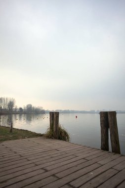Göl kıyısında ahşap demirleme direkleri olan iskele ve İtalya kırsalında bulutlu ve sisli bir günde suya dökülen arka plandaki ağaçlar.