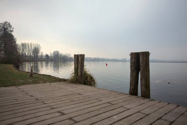 Göl kıyısında ahşap demirleme direkleri olan iskele ve İtalya kırsalında bulutlu ve sisli bir günde suya dökülen arka plandaki ağaçlar.