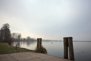 Göl kıyısında ahşap demirleme direkleri olan iskele ve İtalya kırsalında bulutlu ve sisli bir günde suya dökülen arka plandaki ağaçlar.