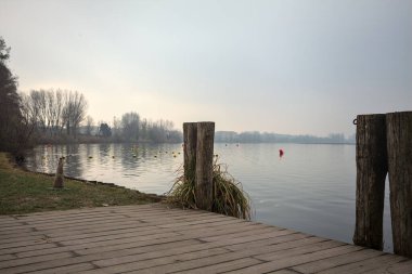 Göl kıyısında ahşap demirleme direkleri olan iskele ve İtalya kırsalında bulutlu ve sisli bir günde suya dökülen arka plandaki ağaçlar.
