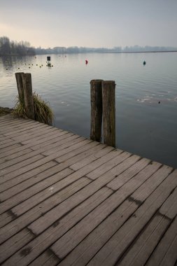 Göl kıyısında ahşap demirleme direkleri olan iskele ve İtalya kırsalında bulutlu ve sisli bir günde suya dökülen arka plandaki ağaçlar.