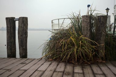 Göl kıyısında ahşap demirleme direkleri olan iskele ve İtalya kırsalında bulutlu ve sisli bir günde suya dökülen arka plandaki ağaçlar.