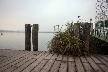Göl kıyısında ahşap demirleme direkleri olan iskele ve İtalya kırsalında bulutlu ve sisli bir günde suya dökülen arka plandaki ağaçlar.