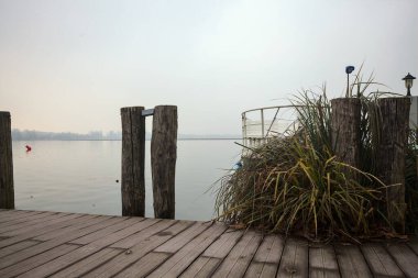 Göl kıyısında ahşap demirleme direkleri olan iskele ve İtalya kırsalında bulutlu ve sisli bir günde suya dökülen arka plandaki ağaçlar.