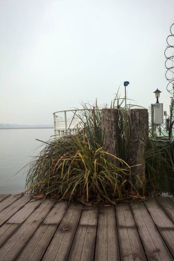 Göl kıyısında ahşap demirleme direkleri olan iskele ve İtalya kırsalında bulutlu ve sisli bir günde suya dökülen arka plandaki ağaçlar.