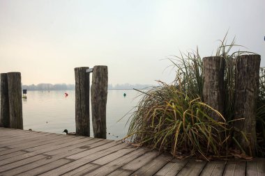 Göl kıyısında ahşap demirleme direkleri olan iskele ve İtalya kırsalında bulutlu ve sisli bir günde suya dökülen arka plandaki ağaçlar.