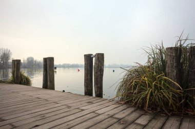 Göl kıyısında ahşap demirleme direkleri olan iskele ve İtalya kırsalında bulutlu ve sisli bir günde suya dökülen arka plandaki ağaçlar.