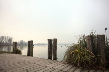 Göl kıyısında ahşap demirleme direkleri olan iskele ve İtalya kırsalında bulutlu ve sisli bir günde suya dökülen arka plandaki ağaçlar.