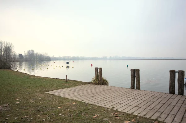 Göl kıyısında ahşap demirleme direkleri olan iskele ve İtalya kırsalında bulutlu ve sisli bir günde suya dökülen arka plandaki ağaçlar.