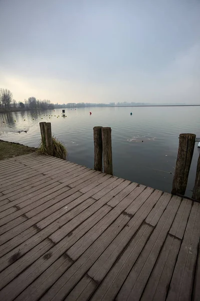 Göl kıyısında ahşap demirleme direkleri olan iskele ve İtalya kırsalında bulutlu ve sisli bir günde suya dökülen arka plandaki ağaçlar.