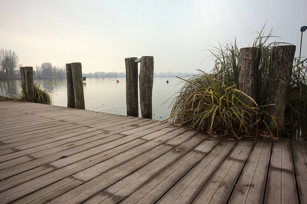 Göl kıyısında ahşap demirleme direkleri olan iskele ve İtalya kırsalında bulutlu ve sisli bir günde suya dökülen arka plandaki ağaçlar.