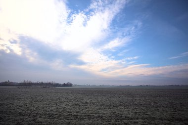 Üzerinde seyrek ağaçlar bulunan, kıpır kıpır tarlalarla kaplı, bir ev silueti ve bir köy kilisesi olan, İtalya 'nın kırsal kesimlerinde bulutlu bir günde