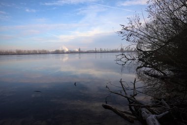 Nehrin karşı kıyısındaki bir ormanın arkasındaki sanayi bölgesinde ağaçlar, binalar ve gökyüzü İtalya kırsalındaki bulutlu bir günde suya yansıyor.