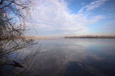 Bulutlu bir günde, İtalya kırsalındaki bir parkta, gökyüzü ve her iki kıyının ağaçlarıyla birlikte suya dökülen bir nehir.