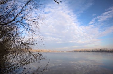 Bulutlu bir günde, İtalya kırsalındaki bir parkta, gökyüzü ve her iki kıyının ağaçlarıyla birlikte suya dökülen bir nehir.