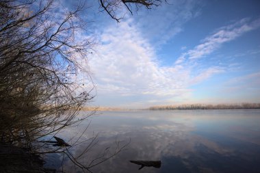 Bulutlu bir günde, İtalya kırsalındaki bir parkta, gökyüzü ve her iki kıyının ağaçlarıyla birlikte suya dökülen bir nehir.