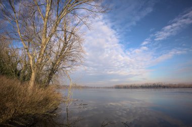 Bulutlu bir günde, İtalya kırsalındaki bir parkta, gökyüzü ve her iki kıyının ağaçlarıyla birlikte suya dökülen bir nehir.