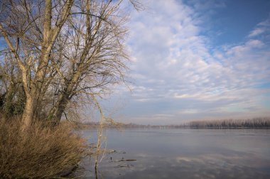 Bulutlu bir günde, İtalya kırsalındaki bir parkta, gökyüzü ve her iki kıyının ağaçlarıyla birlikte suya dökülen bir nehir.