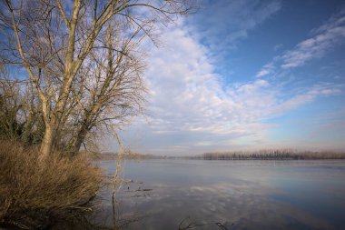 Bulutlu bir günde, İtalya kırsalındaki bir parkta, gökyüzü ve her iki kıyının ağaçlarıyla birlikte suya dökülen bir nehir.