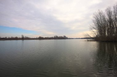 İtalya kırsalında bulutlu bir günde, ormanın çıplak ağaçlarıyla çevrili bir nehir.