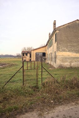 İtalya kırsalında terk edilmiş bir kır evi, bir tarlanın kenarında, giriş kapısının arkasından kısmen açık görünüyor.