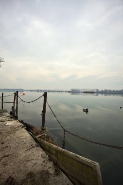 Gölün kıyısındaki bir rıhtımın köşesinde demir bir zincir, yakınlarda yüzen şamandıralar ve kışın İtalya 'nın kırsalında suya düşen bulutlu bir gökyüzü.