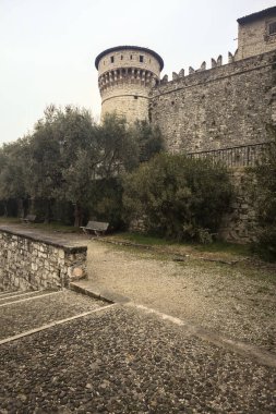 Ağaçlar ve çalılar tarafından çerçevelenmiş, bulutlu bir günde, küçük bir duvarın arkasında, küçük bir duvarın arkasında görülen bir kalenin kalesi.