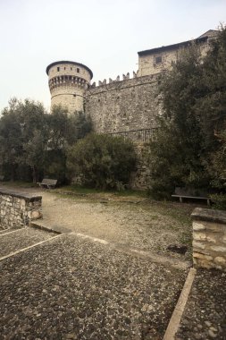 Ağaçlar ve çalılar tarafından çerçevelenmiş, bulutlu bir günde, küçük bir duvarın arkasında, küçük bir duvarın arkasında görülen bir kalenin kalesi.