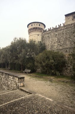 Ağaçlar ve çalılar tarafından çerçevelenmiş, bulutlu bir günde, küçük bir duvarın arkasında, küçük bir duvarın arkasında görülen bir kalenin kalesi.