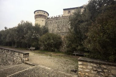 Ağaçlar ve çalılar tarafından çerçevelenmiş, bulutlu bir günde, küçük bir duvarın arkasında, küçük bir duvarın arkasında görülen bir kalenin kalesi.