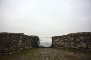 Küçük taş bir duvarın içinde, altında bir şehir, bulutlu ve sisli bir gökyüzünü çerçeveliyor.