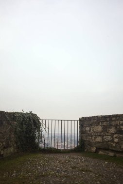 Küçük taş bir duvarın içinde, altında bir şehir, bulutlu ve sisli bir gökyüzünü çerçeveliyor.