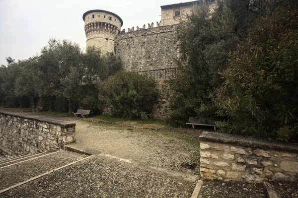 Ağaçlar ve çalılar tarafından çerçevelenmiş, bulutlu bir günde, küçük bir duvarın arkasında, küçük bir duvarın arkasında görülen bir kalenin kalesi.