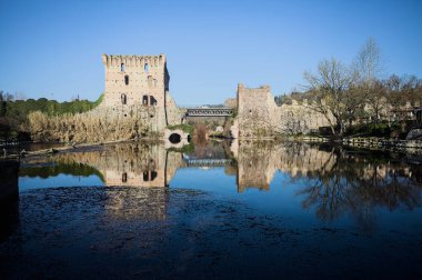 Bir yol köprüsü arasında, içinden geçen ve bir sınır duvarının kalıntıları üzerinde, kışın güneşli bir günde uzaktan görülen yapıyı yansıtan bir nehrin üzerindeki kaleler.