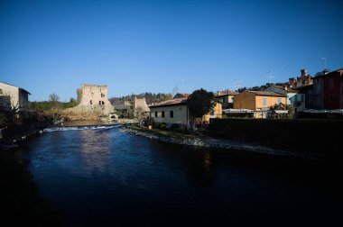 İtalya 'nın kırsal kesimindeki bir köyden geçen nehir, kıyılarında evler olan ve üzerinde kaleler ve eski metalik bir yol köprüsü bulunan bir sınır duvarının kalıntıları olan arka planda.