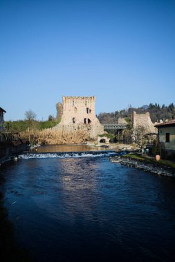 İtalya 'nın kırsal kesimindeki bir köyden geçen nehir, kıyılarında evler olan ve üzerinde kaleler ve eski metalik bir yol köprüsü bulunan bir sınır duvarının kalıntıları olan arka planda.
