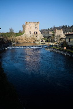 İtalya 'nın kırsal kesimindeki bir köyden geçen nehir, kıyılarında evler olan ve üzerinde kaleler ve eski metalik bir yol köprüsü bulunan bir sınır duvarının kalıntıları olan arka planda.