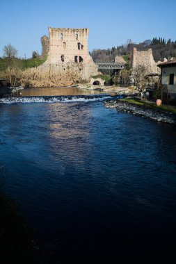 İtalya 'nın kırsal kesimindeki bir köyden geçen nehir, kıyılarında evler olan ve üzerinde kaleler ve eski metalik bir yol köprüsü bulunan bir sınır duvarının kalıntıları olan arka planda.