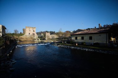 İtalya 'nın kırsal kesimindeki bir köyden geçen nehir, kıyılarında evler olan ve üzerinde kaleler ve eski metalik bir yol köprüsü bulunan bir sınır duvarının kalıntıları olan arka planda.