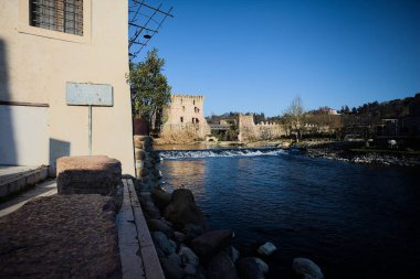 İtalya 'nın kırsal kesimindeki bir köyden geçen nehir, kıyılarında evler olan ve üzerinde kaleler ve eski metalik bir yol köprüsü bulunan bir sınır duvarının kalıntıları olan arka planda.