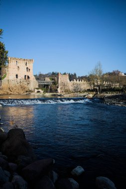 İtalya 'nın kırsal kesimindeki bir köyden geçen nehir, kıyılarında evler olan ve üzerinde kaleler ve eski metalik bir yol köprüsü bulunan bir sınır duvarının kalıntıları olan arka planda.