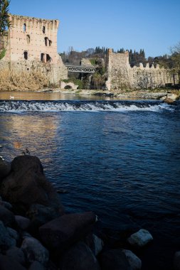 İtalya 'nın kırsal kesimindeki bir köyden geçen nehir, kıyılarında evler olan ve üzerinde kaleler ve eski metalik bir yol köprüsü bulunan bir sınır duvarının kalıntıları olan arka planda.