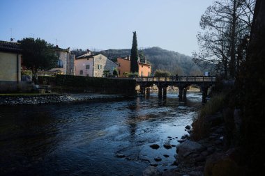 Nehir kıyısındaki bir köyün yanında akan bir nehrin üzerinde, güneşli bir günde görülen taş ve ahşap bir köprü.