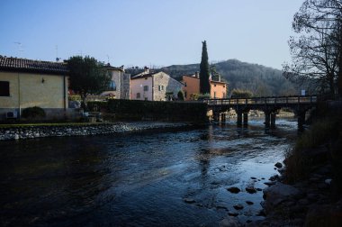 Nehir kıyısındaki bir köyün yanında akan bir nehrin üzerinde, güneşli bir günde görülen taş ve ahşap bir köprü.