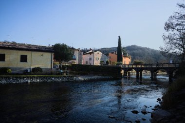 Nehir kıyısındaki bir köyün yanında akan bir nehrin üzerinde, güneşli bir günde görülen taş ve ahşap bir köprü.