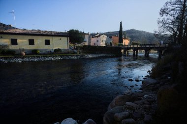 Nehir kıyısındaki bir köyün yanında akan bir nehrin üzerinde, güneşli bir günde görülen taş ve ahşap bir köprü.